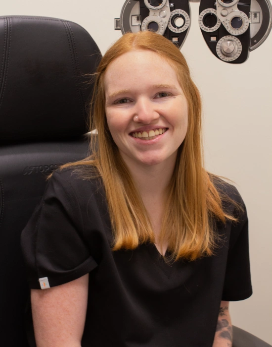 A woman with long red hair, wearing black scrubs, sits and smiles in an exam room with ophthalmology equipment visible in the background, reflecting her role as a trusted Twin Falls eye doctor.
