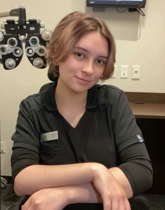 A woman wearing black scrubs sits at a desk in an eye clinic, with optometry equipment and a TV on the wall in the background.