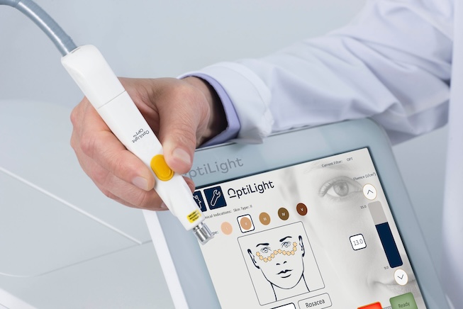 A person holds a medical device above a touchscreen displaying OptiLight settings and a facial diagram, demonstrating advanced Eye Care for precise Dry Eye Treatment guidance.