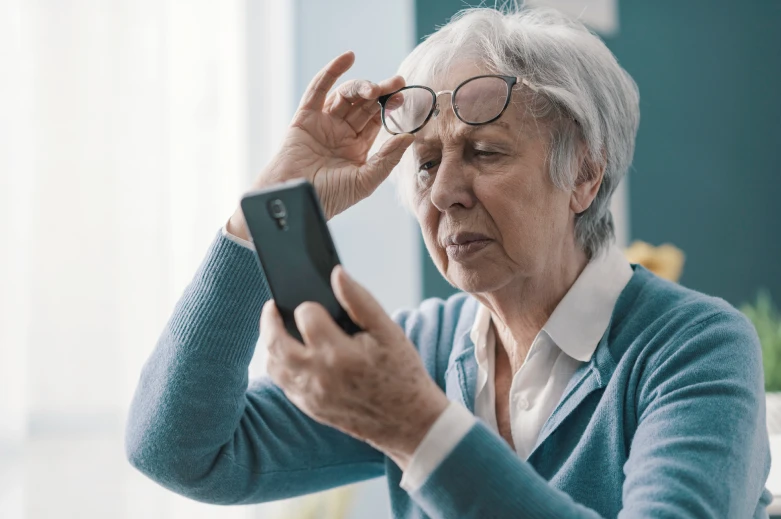 Older woman holding her glasses above her eyes and squinting at a smartphone, possibly experiencing difficulty reading due to Macular Degeneration.