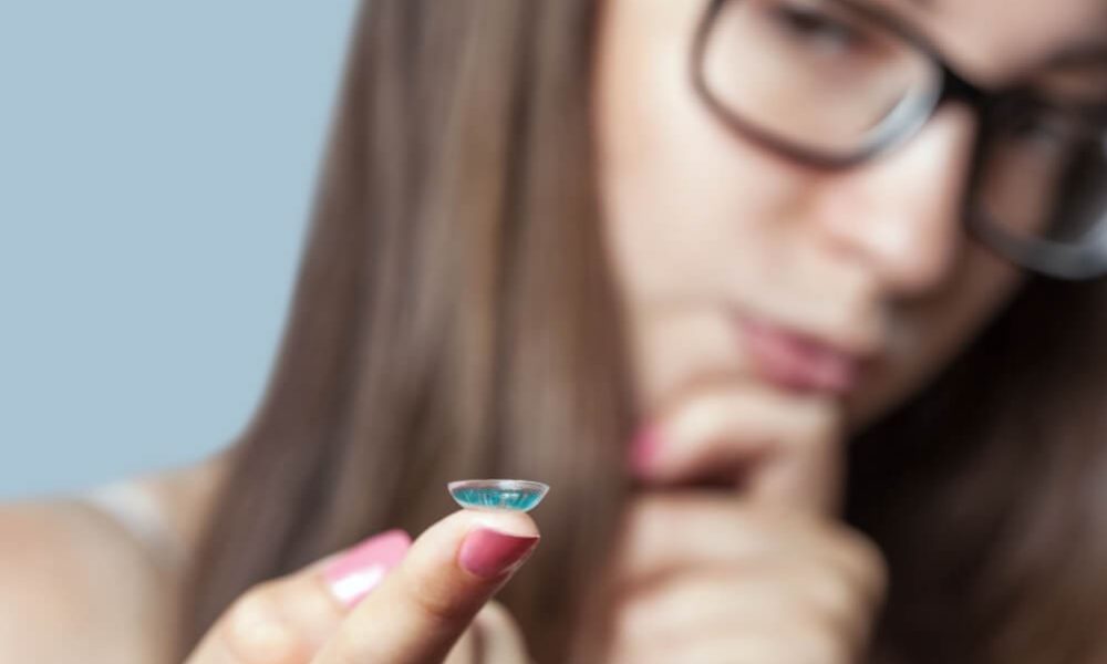 A woman holding a small blue colored contact lens.