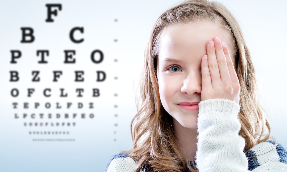 A girl covers one eye with her hand while standing in front of an eye chart, hoping to see clearly despite her blurry vision.