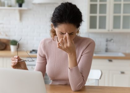 A woman sitting at a table in a kitchen holds her glasses in one hand and pinches the bridge of her nose with the other, struggling to see clearly while working on a laptop.