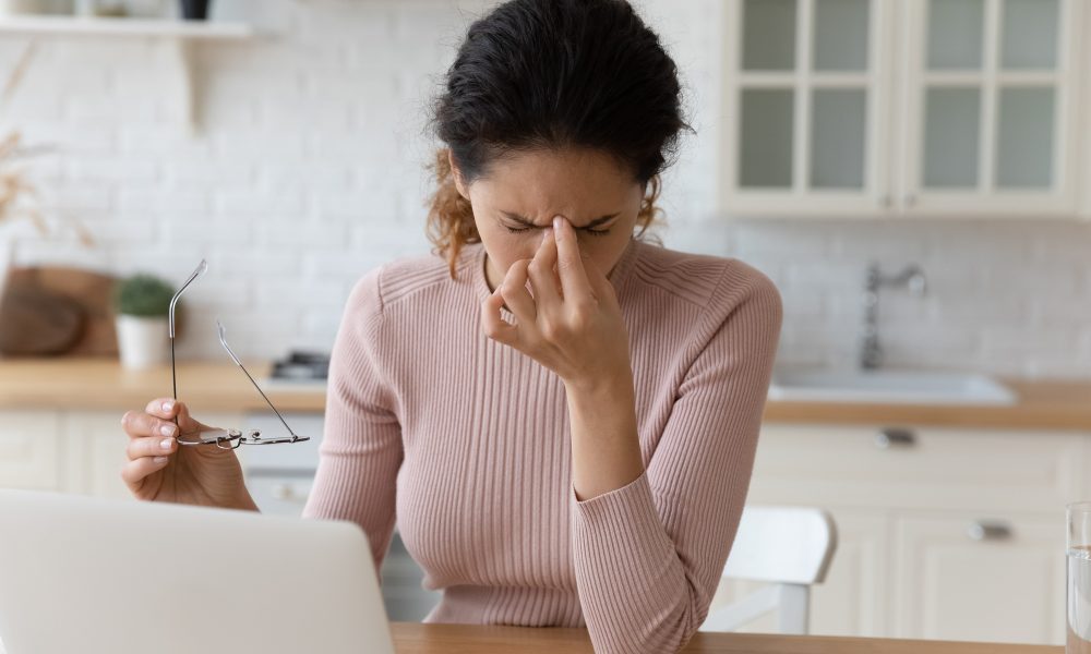 A woman sitting at a table in a kitchen holds her glasses in one hand and pinches the bridge of her nose with the other, struggling to see clearly while working on a laptop.