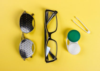 Flat lay of eyeglasses, pinhole glasses, and a contact lens case with tweezers on a vibrant yellow background, showcasing vision care essentials.