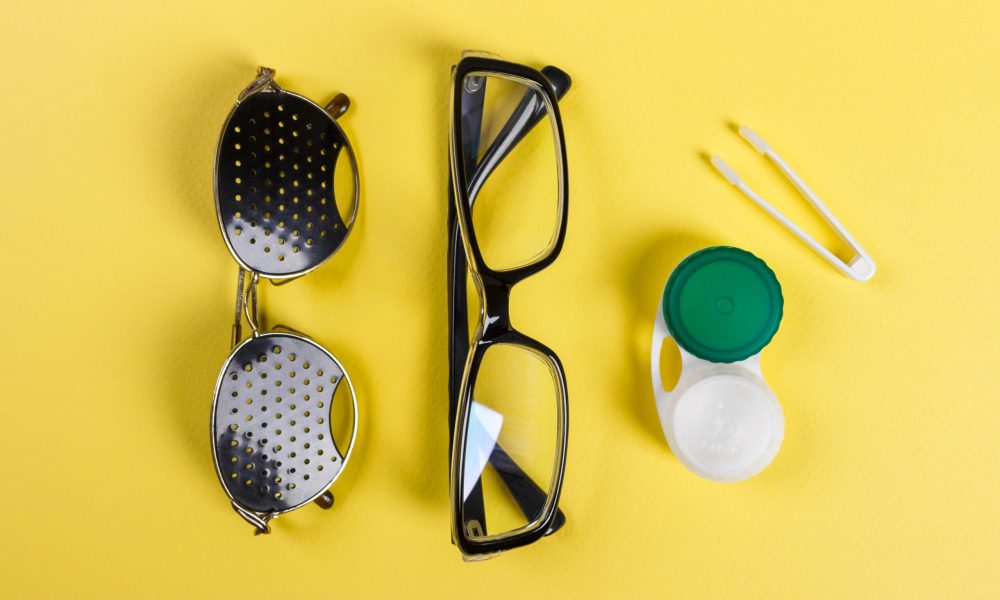 Flat lay of eyeglasses, pinhole glasses, and a contact lens case with tweezers on a vibrant yellow background, showcasing vision care essentials.