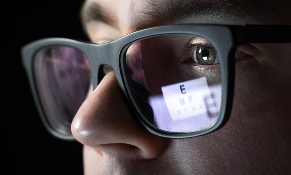 Close-up of a person wearing glasses, with an eye chart visible in the reflection of the lenses.