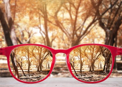 Red glasses rest on a surface, making the trees in the background appear clearer and more vibrant through the lenses, contrasting sharply with the blurred scenery beyond. According to eye experts, this effect can bring any environment to life, even in climates as varied as Boise's.