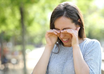 A woman in a gray sweater rubs her eyes outdoors on a sunny day, enjoying the vibrant surroundings. Maintaining healthy eyes is vital to prevent eye infections, especially when enjoying nature's beauty with a blurred green backdrop.