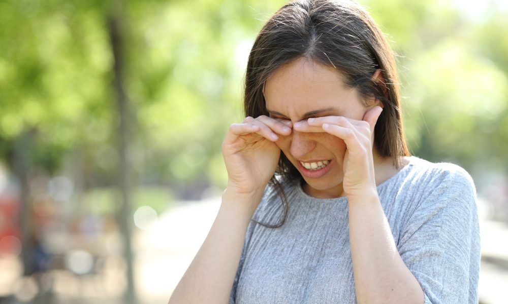 A woman in a gray sweater rubs her eyes outdoors on a sunny day, enjoying the vibrant surroundings. Maintaining healthy eyes is vital to prevent eye infections, especially when enjoying nature's beauty with a blurred green backdrop.