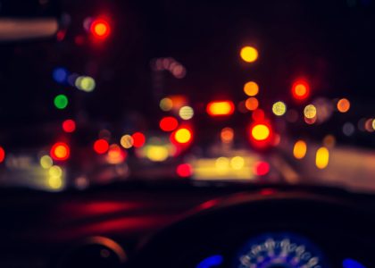 The blurred view from inside a vehicle at night, with unfocused traffic and car lights ahead, captures the challenge of night driving for those with astigmatism. A dashboard sits partially visible in the foreground, adding to this hazy yet familiar scene.