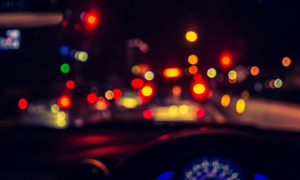 The blurred view from inside a vehicle at night, with unfocused traffic and car lights ahead, captures the challenge of night driving for those with astigmatism. A dashboard sits partially visible in the foreground, adding to this hazy yet familiar scene.