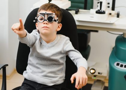 A young boy sits in an eye examination chair at Logan's Eye Specialists, wearing trial frames and pointing forward, with ophthalmology equipment neatly arranged in the background.