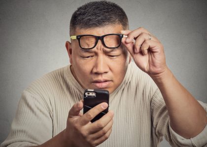 A man squints while holding his smartphone close, adjusting his glasses as he looks intently at the screen. Sporting a light-colored sweater against a plain background, he's focused on correcting vision issues, balancing monovision for both near and far vision tasks.