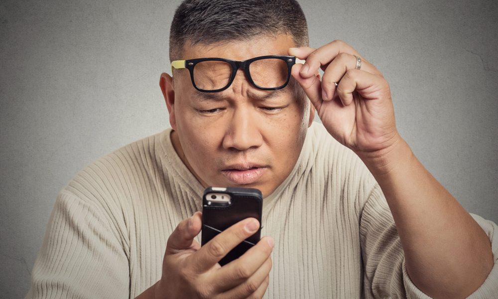 A man squints while holding his smartphone close, adjusting his glasses as he looks intently at the screen. Sporting a light-colored sweater against a plain background, he's focused on correcting vision issues, balancing monovision for both near and far vision tasks.