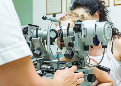 An eye doctor examines a child's vision using a phoropter, with the child sitting on an adult's lap in an optometry office.