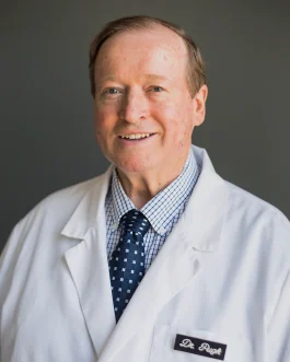 A Boise eye doctor in a white medical coat and a dark tie stands against a plain background, smiling slightly.