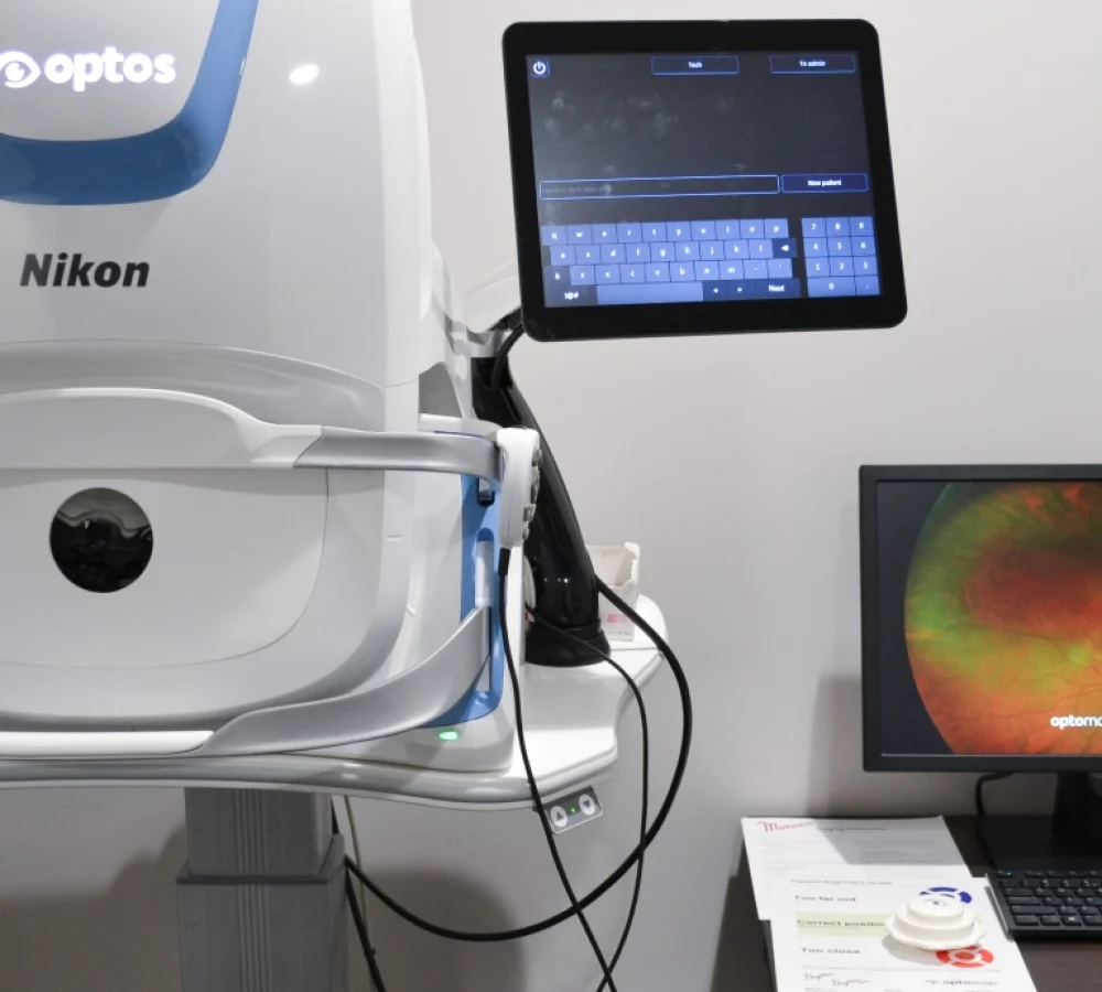 A Nikon Optos eye imaging machine with an attached touchscreen, a computer monitor displaying an eye scan, and paperwork on a nearby table in a Spanish Fork eye doctor’s office.