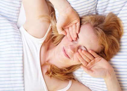 A woman laying in bed with her contacts in, her hands covering her eyes.