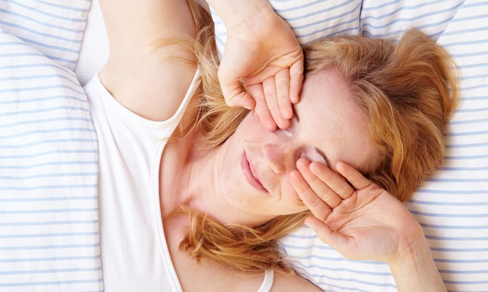 A woman laying in bed with her contacts in, her hands covering her eyes.