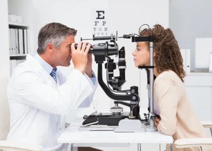 An eye doctor examines a woman’s eyes with a slit lamp in a clinical office, with an eye chart visible in the background—offering same-day eye exams in Coeur d’Alene.