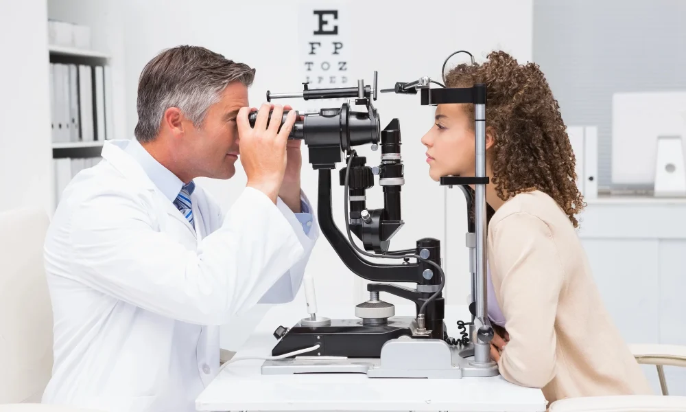 An eye doctor examines a woman’s eyes with a slit lamp in a clinical office, with an eye chart visible in the background—offering same-day eye exams in Coeur d’Alene.