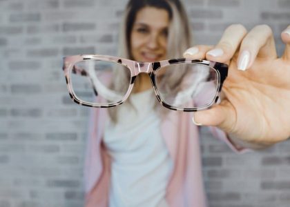 A woman holding a pair of glasses in front of a brick wall, contemplating how often she should get her eyes checked.