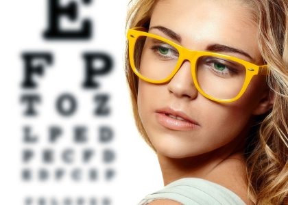 A young woman wearing glasses stands in front of an eye chart, highlighting the importance of detecting eye problems early with optometrists.