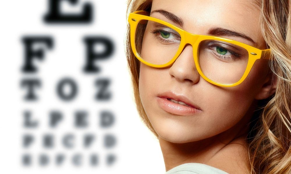 A young woman wearing glasses stands in front of an eye chart, highlighting the importance of detecting eye problems early with optometrists.