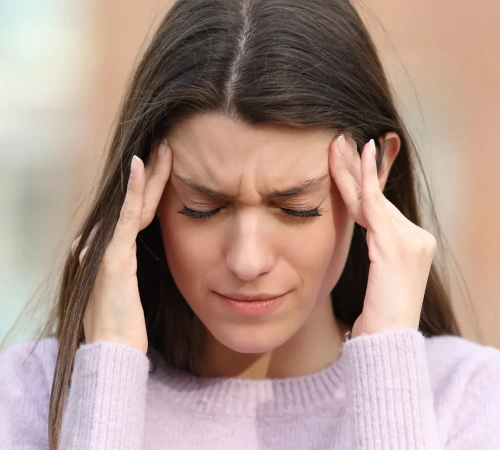 A woman with long brown hair holds her temples and closes her eyes, appearing to have a headache or stress, while wearing a light purple sweater—possibly seeking Dry Eye Relief.